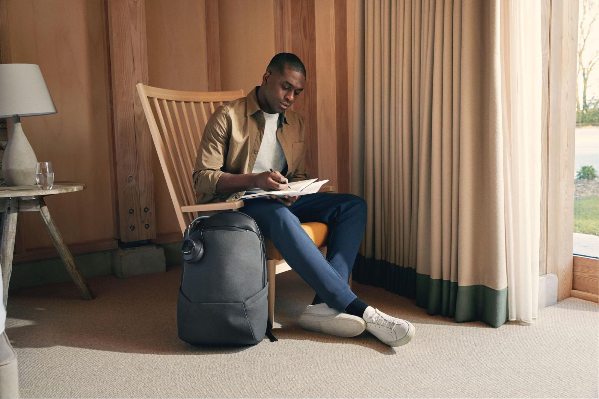  A man seated in a wooden chair writing in a journal is next to a Troubadour brand backpack on the floor.