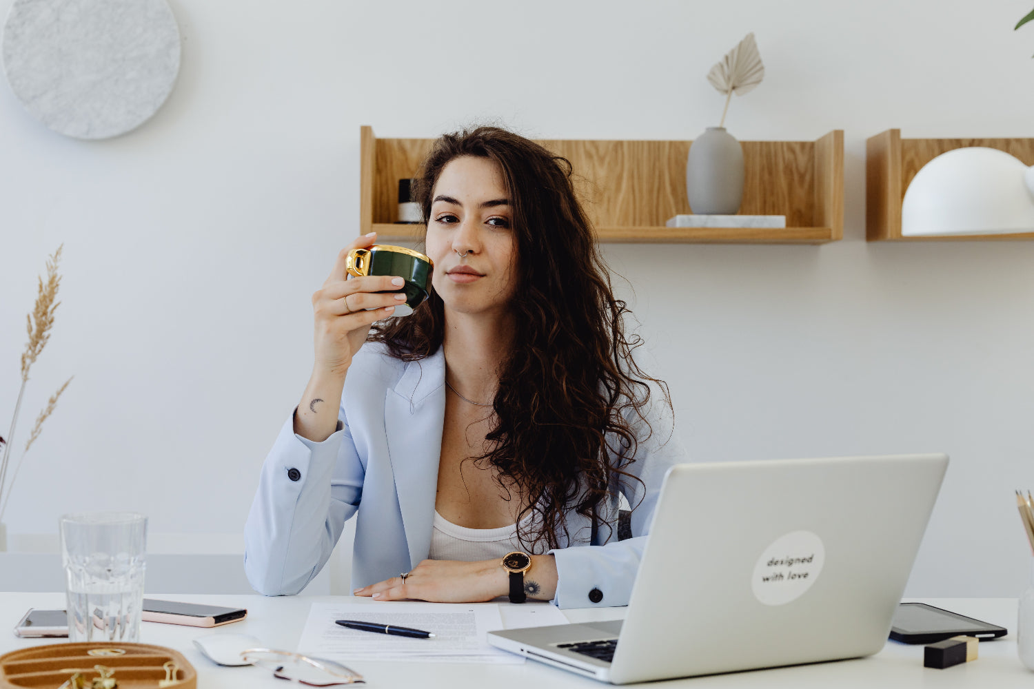 A woman drinking from a coffee mug sitting at a desk in front of an open laptop.