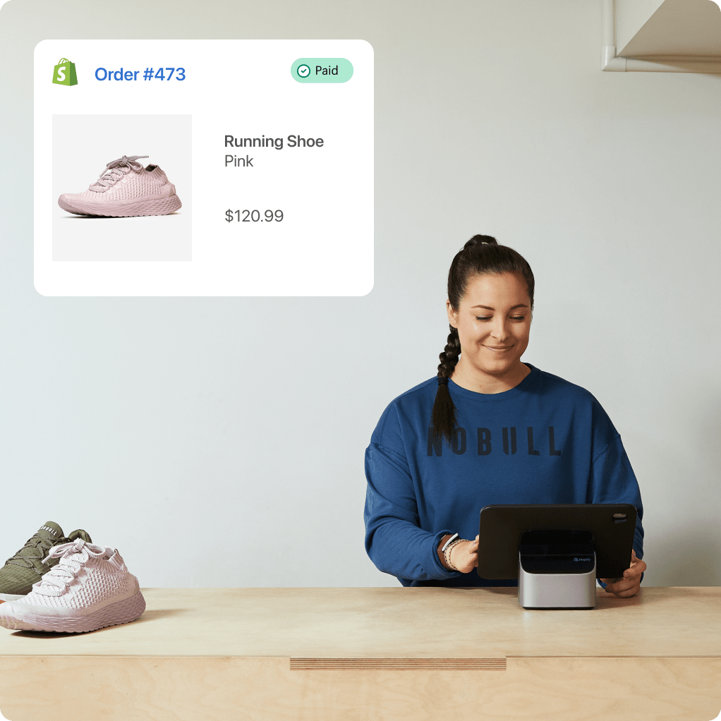 A smiling woman sits at a counter, smiling at a tablet device in front of her. There are two shoes to the left of her, and in the upper left, an order paid confirmation screen with a shoe, a description of “Running shoe - pink” and a cost of $160.60.