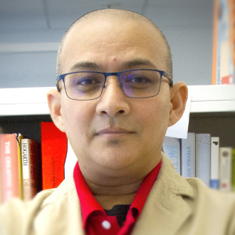 Binod is facing forward. He is bald, wears glasses, a red shirt and a yellow jacket.