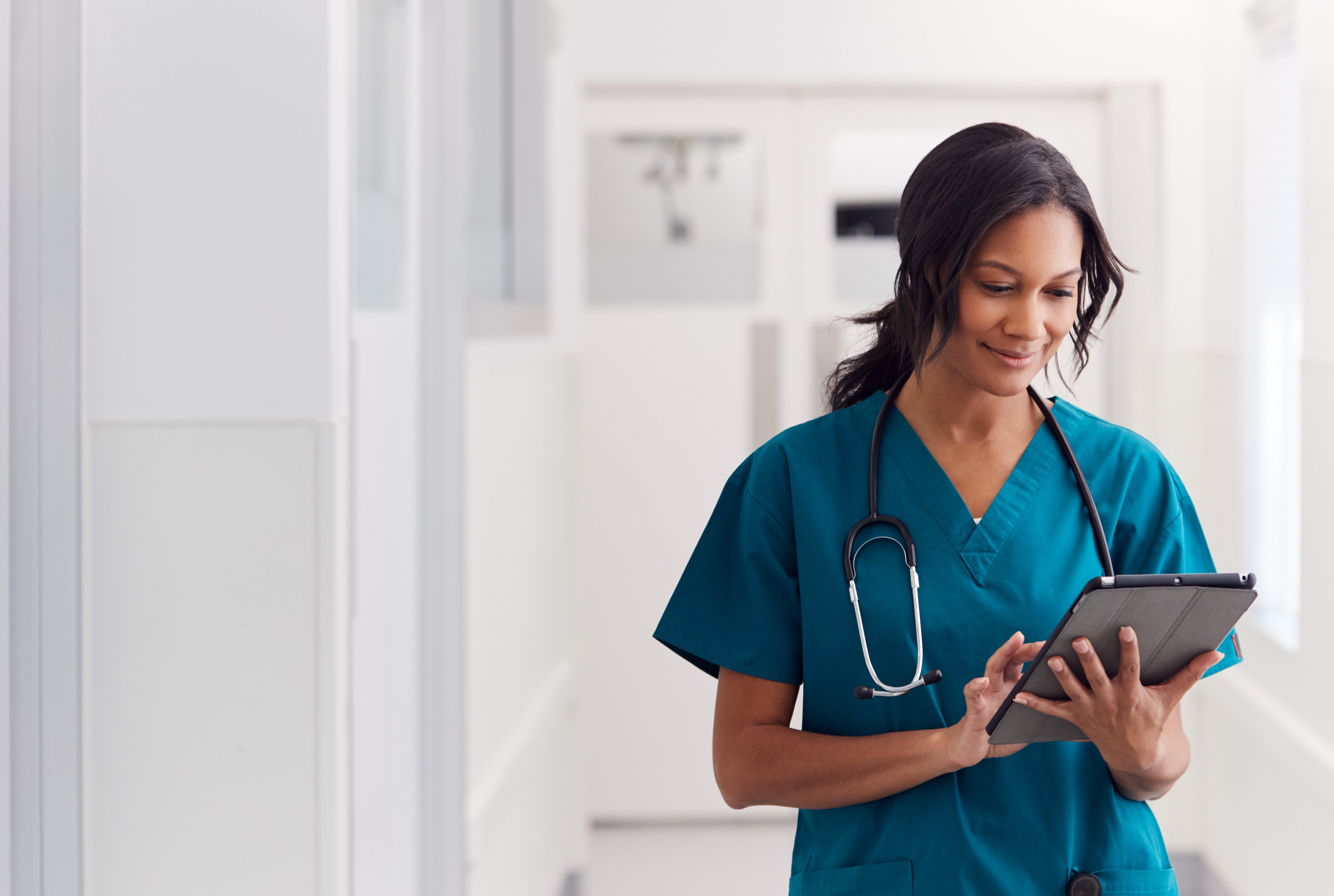 Female Doctor Wearing Scrubs In Hospital Corridor Using Digital Tablet