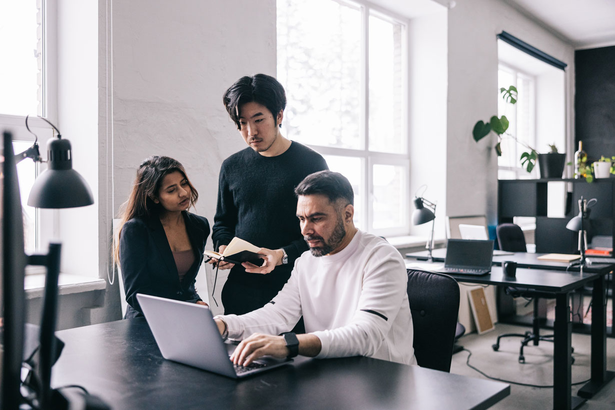 man sitting looking at a laptop with a woman and man standing behind him