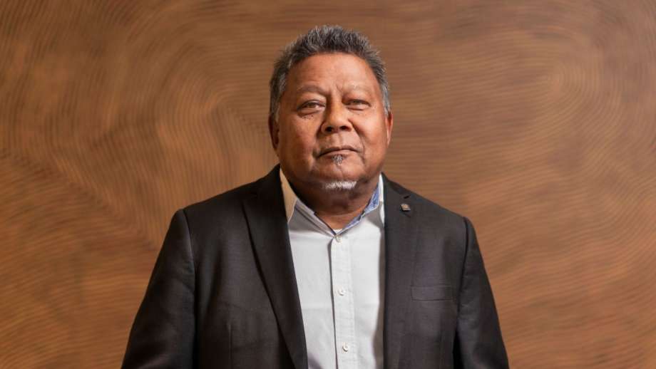 A man wearing a black suit jacket stands in front of an ochre-coloured painting. 