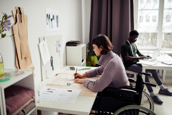 Creative young woman sitting in wheelchair at her desk in studio and drawing sewing pattern