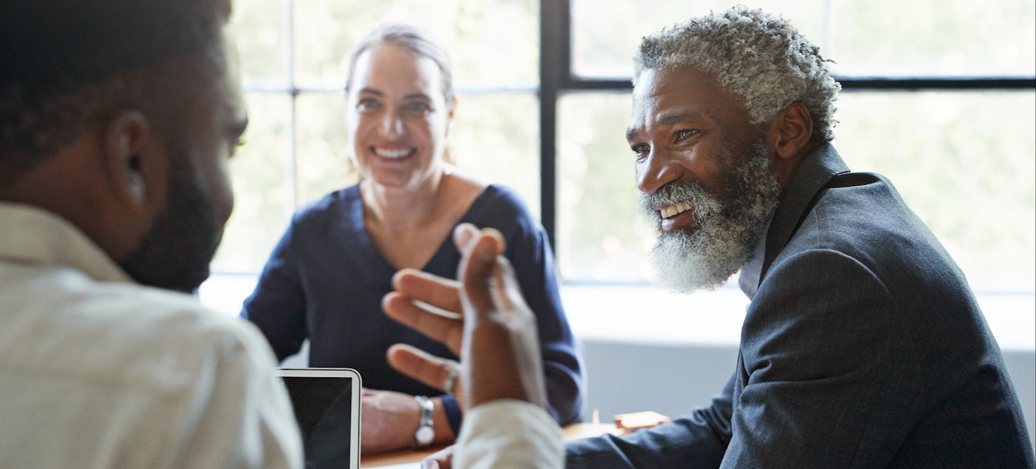 [Featured image] Two men and a woman meet at a conference table with an office window in the background.