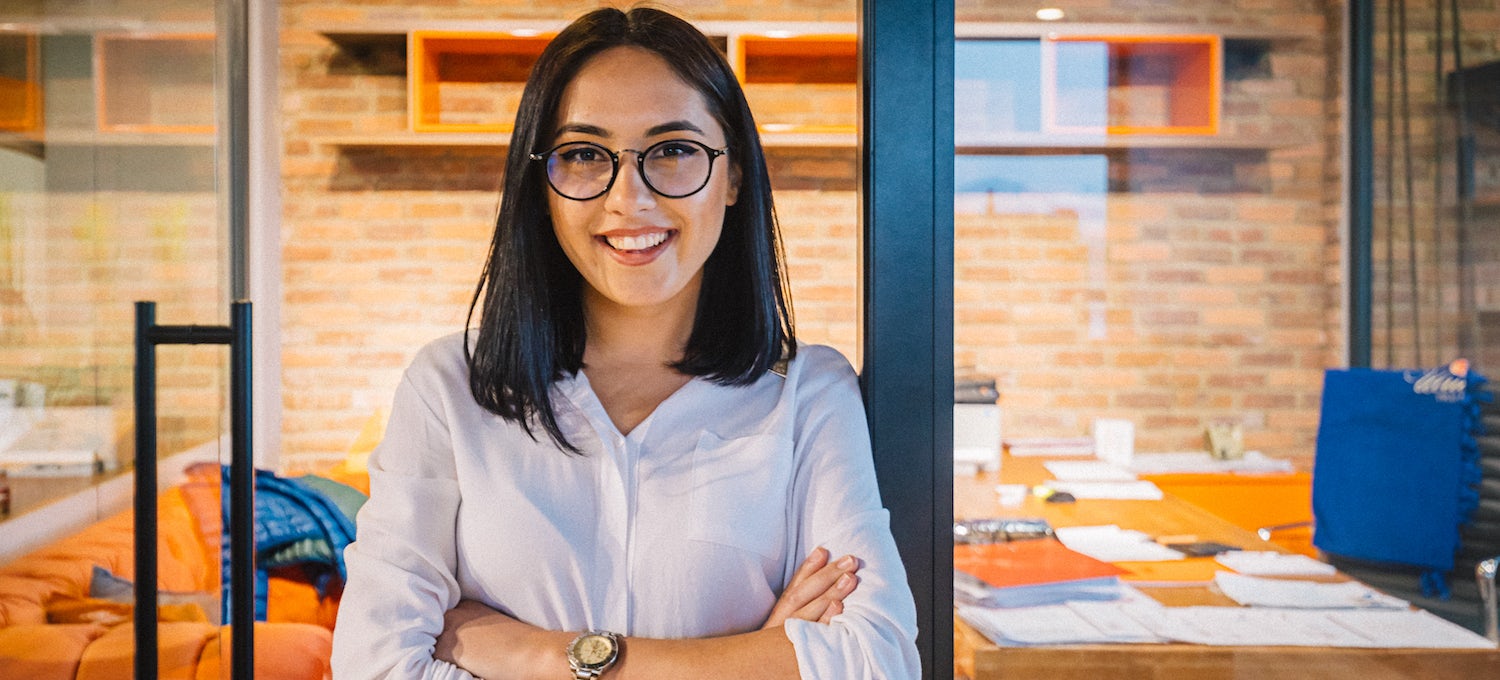 [Featured image] A young graduate student wearing a white button-up shirt and glasses stands with both arms crossed in front of an office. 