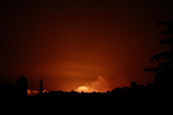 Color photograph of eruption glow at night and the silhouette of trees in the foreground