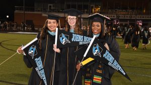 Three people in graduation regalia smiling and holding Johns Hopkins University flags.