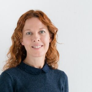 A woman with wavy, shoulder-length red hair smiles gently at the camera. She is wearing a navy blue sweater with a Peter Pan collar. The background is plain and white.