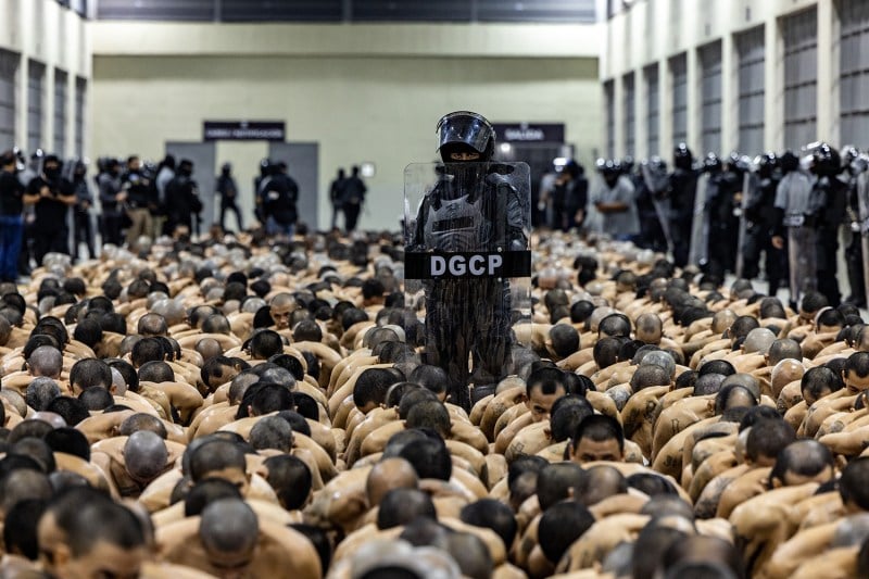 Hundreds of prisoners are lined up in rows, kneeling on the floor with a prison guard standing in the middle, dressed in a black uniform and holding up a clear riot shield.