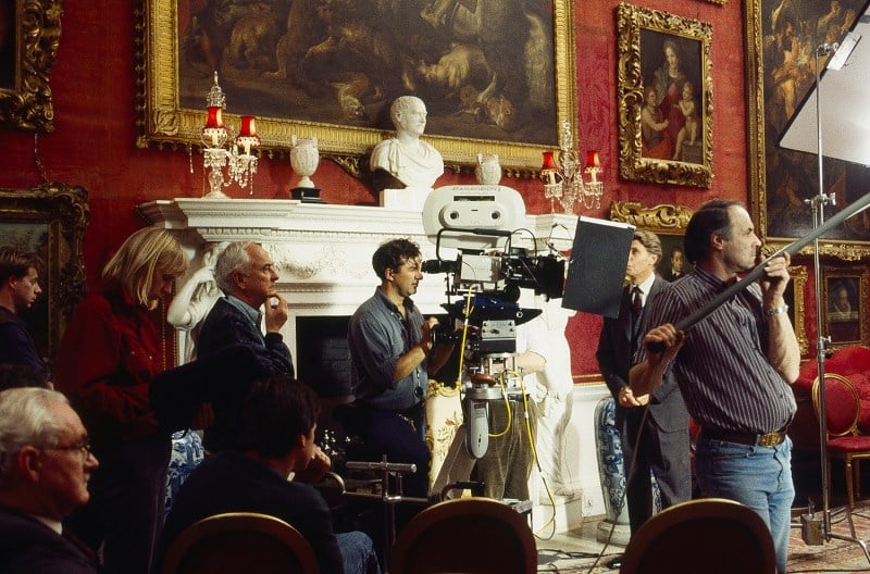 Filmmakers, one positioned behind a camera, stand in an opulent room with paintings, a mantle and bust, while making a movie.