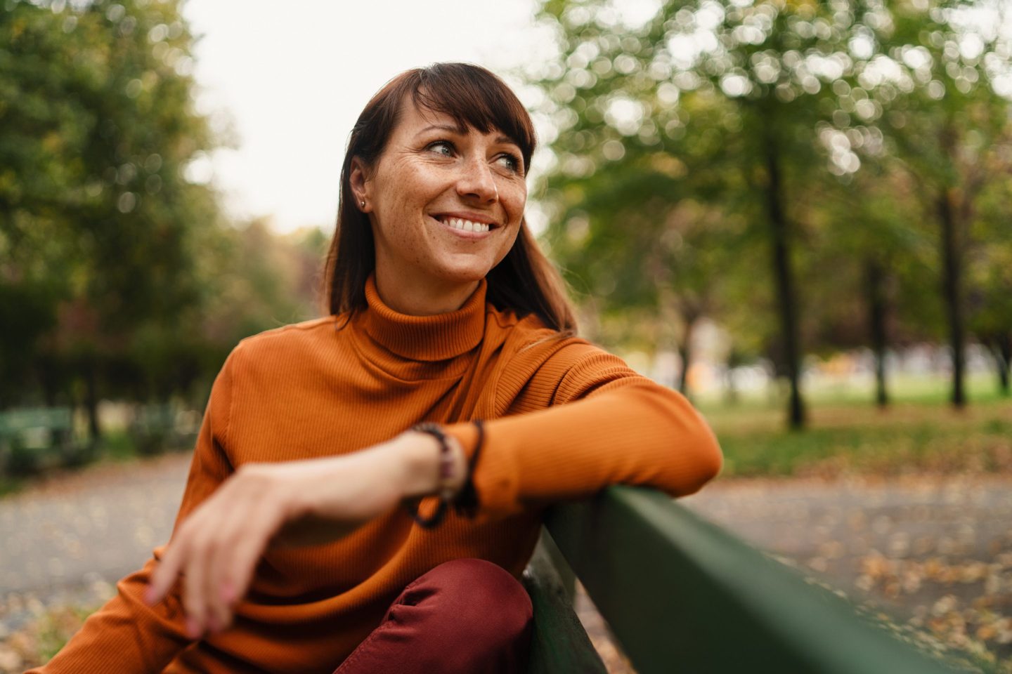 Beautiful mid adult woman sitting on bench and resting outside in the city on autumn day.