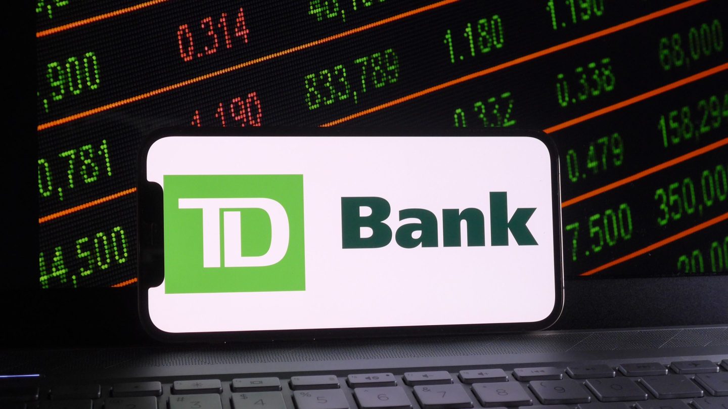 In this photo illustration, Toronto-Dominion Bank logo is displayed on a smartphone with a stock market and a laptop keyboard background.