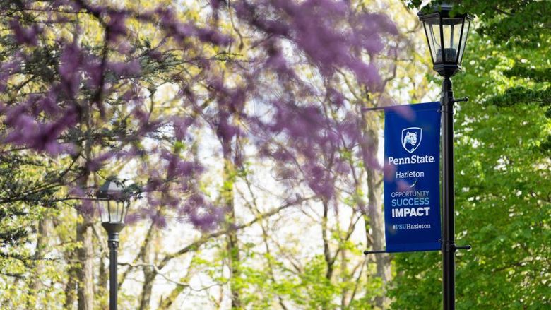 A blue Penn State Hazleton banner on a lightpost with a pink flowered tree in the foreground.