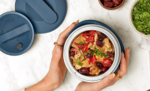 A person holds a round, stainless steel food container with a navy blue lid. The container is filled with a cooked meal of chicken, olives, and vegetables.  A matching lid is placed next to the container on a marble-like countertop.