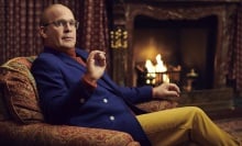 A man in a blue suit jacket, red turtleneck, and yellow pants sits in an armchair by a fireplace.