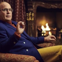 A man in a blue suit jacket, red turtleneck, and yellow pants sits in an armchair by a fireplace.