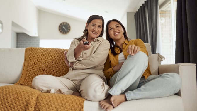 two people sit on a cream-colored couch while one of them hold a TV remote and both smile