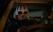 A woman drives a car wearing sunglasses and a sequined hat.