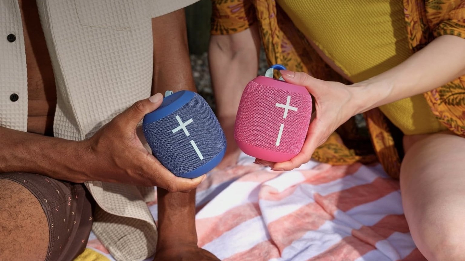 two people holding blue and pink ultimate ears wonderboom 4 spoeakers while sitting on towel outside