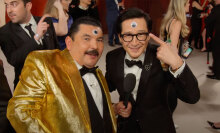 A man in a tuxedo stands next to a man in a gold tuxedo on the red carpet.