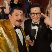 A man in a tuxedo stands next to a man in a gold tuxedo on the red carpet.