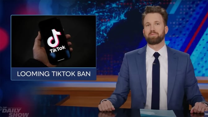 A man sits behind a talk show desk with an image of the TikTok logo on the left.