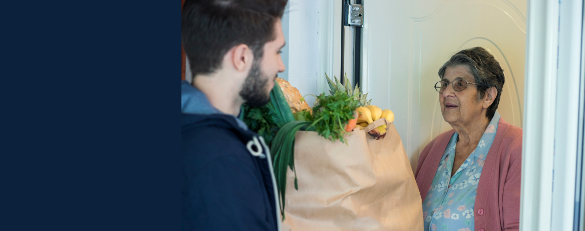 A man holds a bag of groceries out to an elderly woman at her front door.