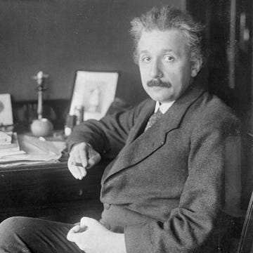 albert einstein sitting at a desk, wearing a suit and tie, looking directly at the camera