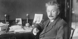 albert einstein sitting at a desk, wearing a suit and tie, looking directly at the camera