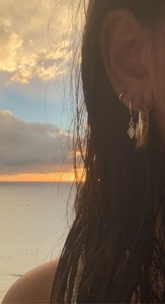 a woman with long hair and earrings standing on the beach looking out at the ocean