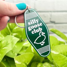 a person holding a keychain that says silly goose club in front of some green plants