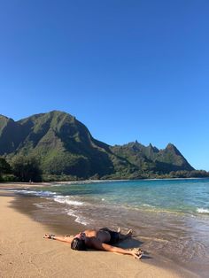 a woman laying on top of a sandy beach next to the ocean with mountains in the background