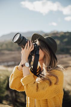 a woman wearing a hat and holding a camera up to her face while looking into the distance