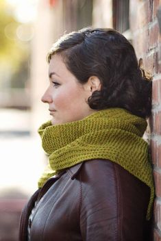 a woman leaning against a brick wall wearing a green knitted cowl neck scarf