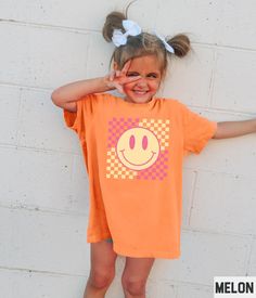 Our retro smiley face graphic tee features a happy face and checkered background design. This tshirt pairs perfectly with the adult, youth, and toddler girls versions (linked below). ✩ ✩ 𝗧𝗼𝗱𝗱𝗹𝗲𝗿 𝗕𝗲𝗹𝗹𝗮 𝗖𝗮𝗻𝘃𝗮𝘀 𝗧𝗲𝗲: https://starrieapparel.etsy.com/listing/1703012396 ✩ 𝗞𝗶𝗱𝘀 𝗕𝗲𝗹𝗹𝗮 𝗖𝗮𝗻𝘃𝗮𝘀 𝗧𝗲𝗲: https://starrieapparel.etsy.com/listing/1702998672 ✩ 𝗞𝗶𝗱𝘀 𝗖𝗼𝗺𝗳𝗼𝗿𝘁 𝗖𝗼𝗹𝗼𝗿𝘀 𝗧𝗲𝗲: https://starrieapparel.etsy.com/listing/1728079245 ✩ 𝗔𝗱𝘂𝗹𝘁 𝗕𝗲𝗹𝗹𝗮 Affordable Short Sleeve T-shirt With Smiley Face, Cheap Smiley Face Short Sleeve T-shirt, Cheap Funny T-shirt With Smiley Face, Fun Smiley Face Short Sleeve T-shirt, Cute Short Sleeve Top With Smiley Face, Pink Short Sleeve T-shirt With Smiley Face, Cute Smiley Face Short Sleeve T-shirt, Trendy Short Sleeve T-shirt With Smiley Face, Playful Orange Graphic Print Tops