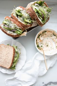 two sandwiches with lettuce, cheese and other toppings sit on plates next to a bowl of dip