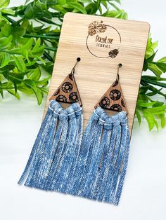 two pairs of wooden earrings with tassels and blue fabric on the bottom, sitting next to green leaves