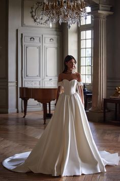 a woman standing in front of a chandelier wearing a wedding dress with an off the shoulder neckline