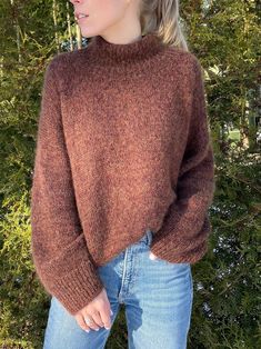 a woman wearing a brown sweater and jeans standing in front of some trees with her hands on her hips