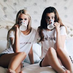 two beautiful young women sitting on top of a bed drinking from mugs in front of their faces