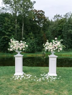 two white pedestals with flowers on them in front of a pond and trees,