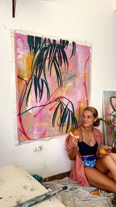 a woman sitting on the floor in front of a painting and eating something with her hand