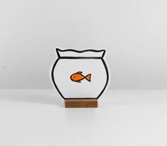 an orange fish in a black and white bowl on a wooden stand with a white wall behind it