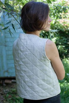 a woman wearing a white quilted vest and black jeans standing in front of trees