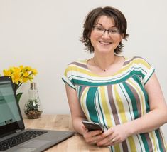 a woman standing in front of a laptop computer holding a cell phone and looking at the camera