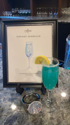 there is a blue and green drink on the table next to a framed sign that says tiffany sparkler