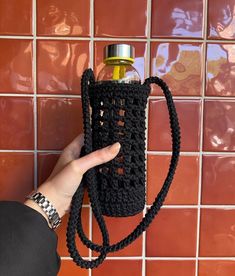 a hand holding a black crocheted water bottle holder in front of a tiled wall