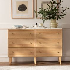 a wooden dresser sitting next to a plant in a room with pictures on the wall