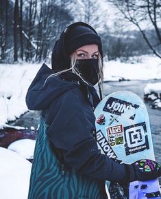 a woman holding a snowboard standing in the snow next to a river and trees